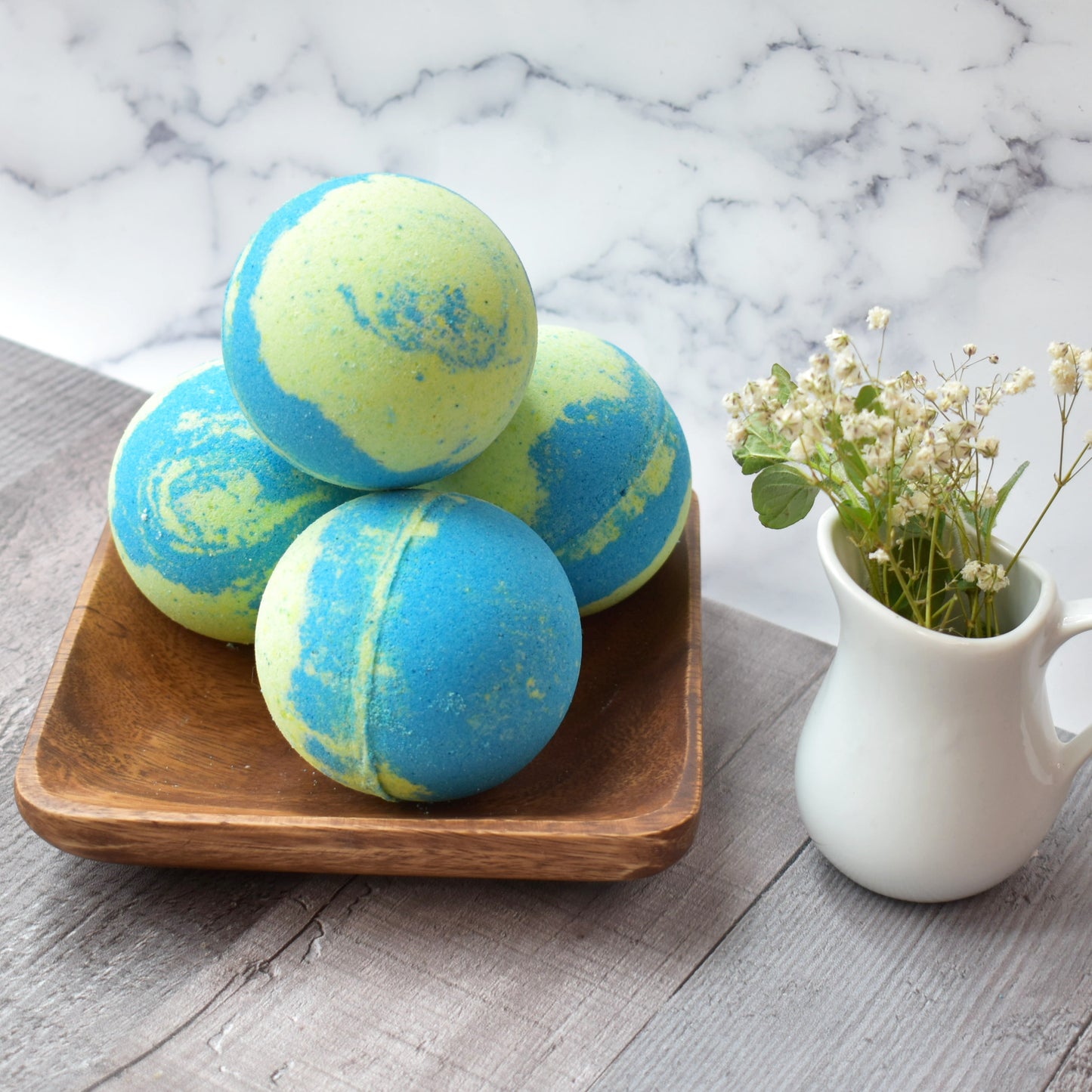 4 Lime green and blue swirl bath bombs are stacked on a square wood tray. Next to bath bombs is a white creamer carafe with baby’s breath sitting on a grey/brown wood table with a marbled background