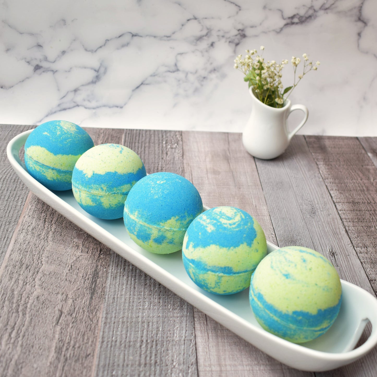 5 Green & Blue swirled bath bombs on a white tray. Small white creamer carafe with baby’s breath are in background Sitting on a light brown/grey wood table and marble backboard