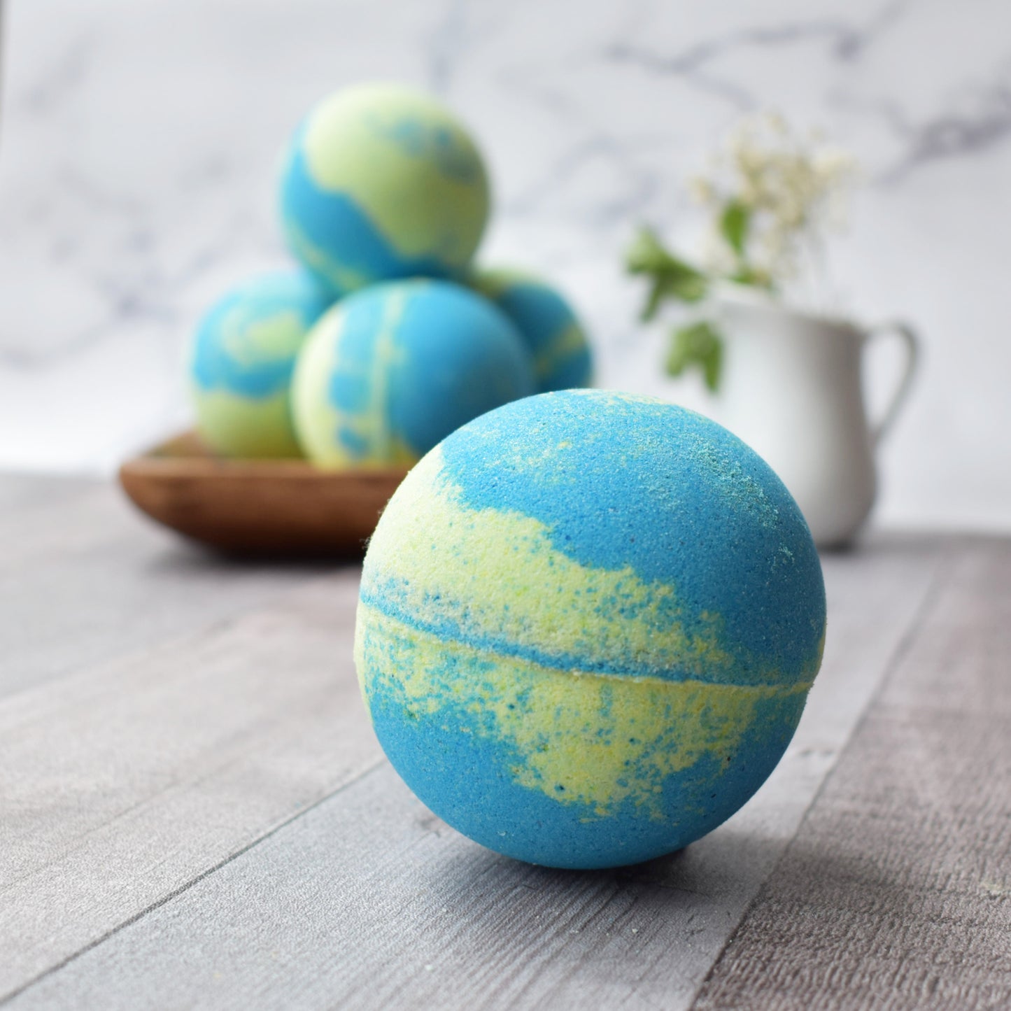 Close up of a Lime green and blue swirled bath bomb. In the background are 4 bath bombs stacked on a square wood tray. The the right is a small white creamer carafe with baby’s breath flowers. All sitting on a wood brown/grey table and marble background