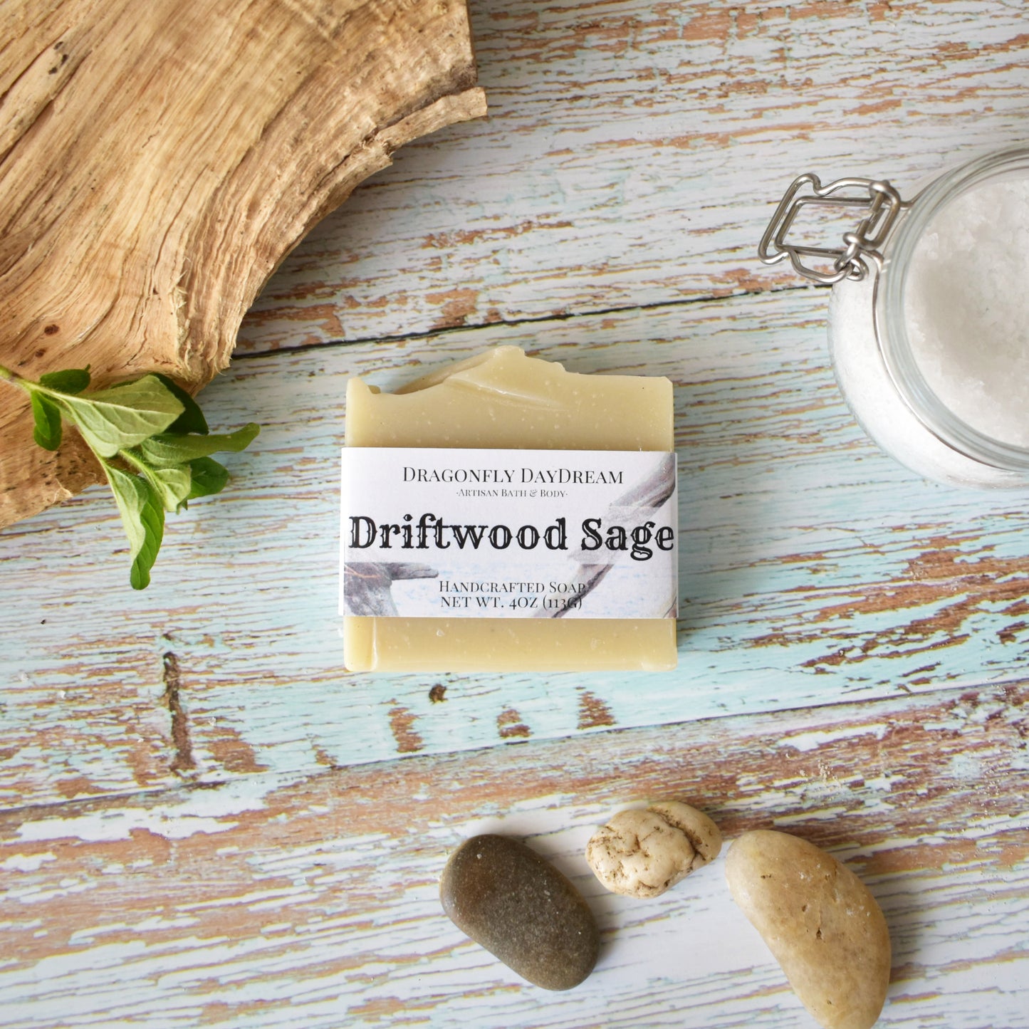 1 labeled rectangular beige soap “Driftwood Sage, Handcrafted Soap, Net wt. 4oz (113g), Dragonfly DayDream, Artisan Bath & Body. Soap is on weathered light blue wood background. To the right of the soap is a clear jar of course white salt, to the left is a piece of driftwood with greenery and to the bottom are three smooth river stones, two light beige and one dark grey.