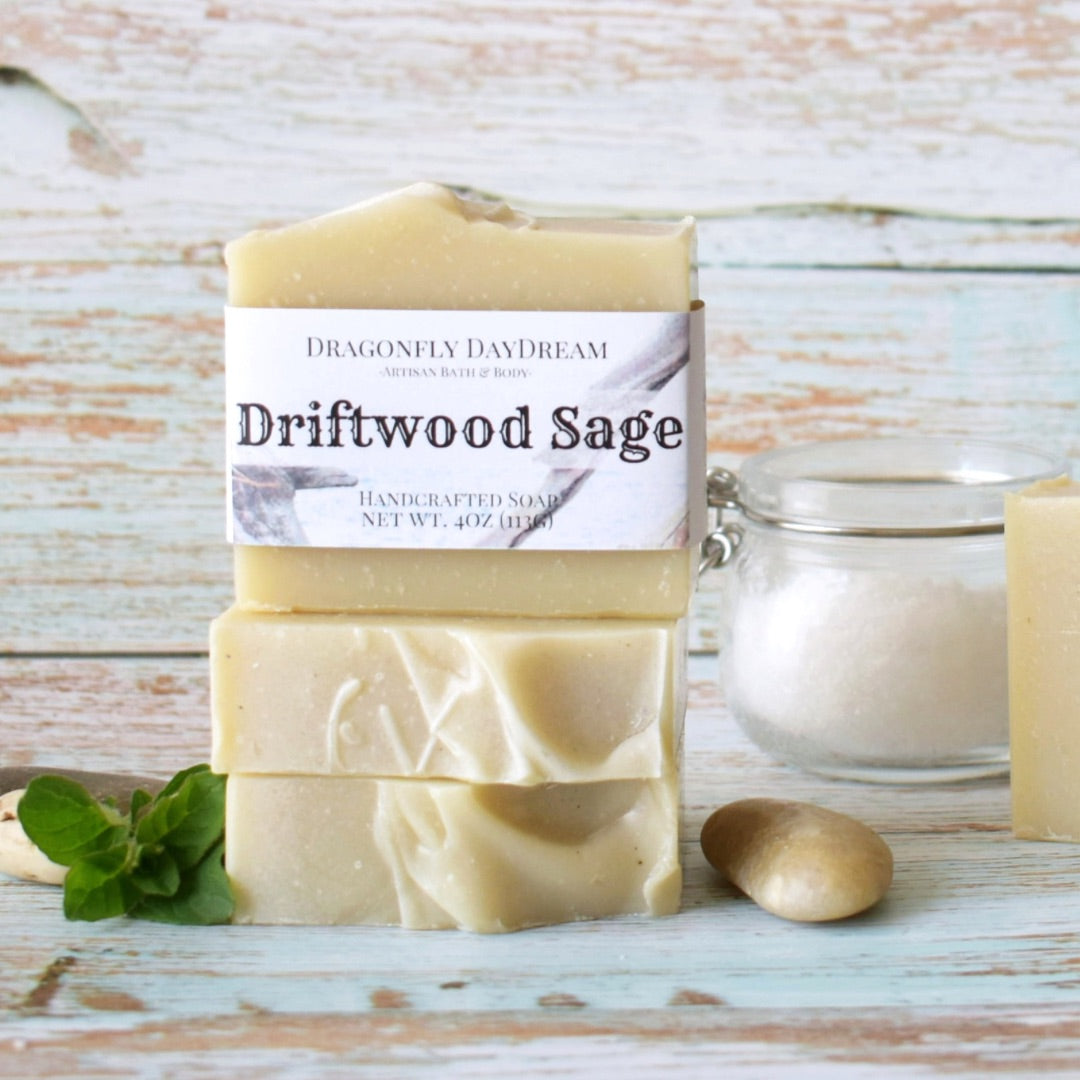 Driftwood Sage Artisan Soap Stacked. Light beige soaps stacked against a light blue wood background with small pebble and jar of salt as props.