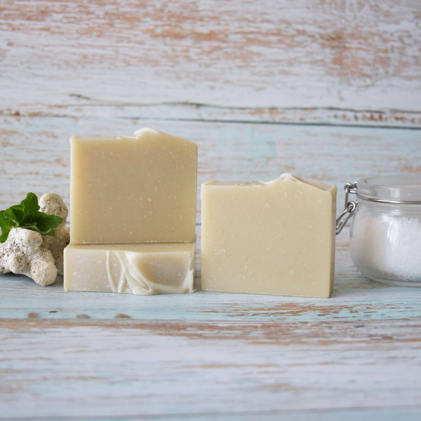 3 beige soaps sitting on a weathered light blue wood background. To the left of the soap is white coral with greenery and to the left is a lightening sealed jar with course salt.