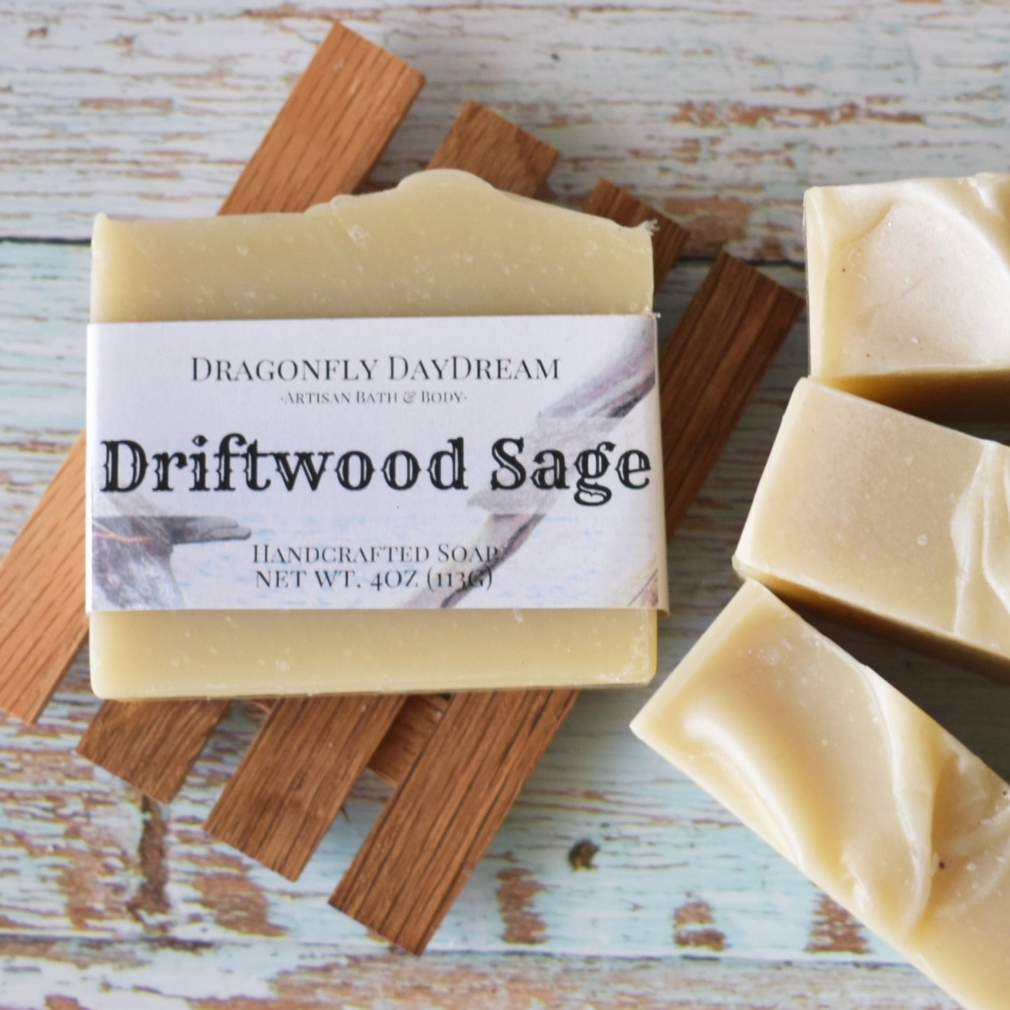Driftwood Sage Artisan Soap Bar sitting on top of a slatted wooden soap tray. To the right are 3 additional soaps with a blueish weathered wood background