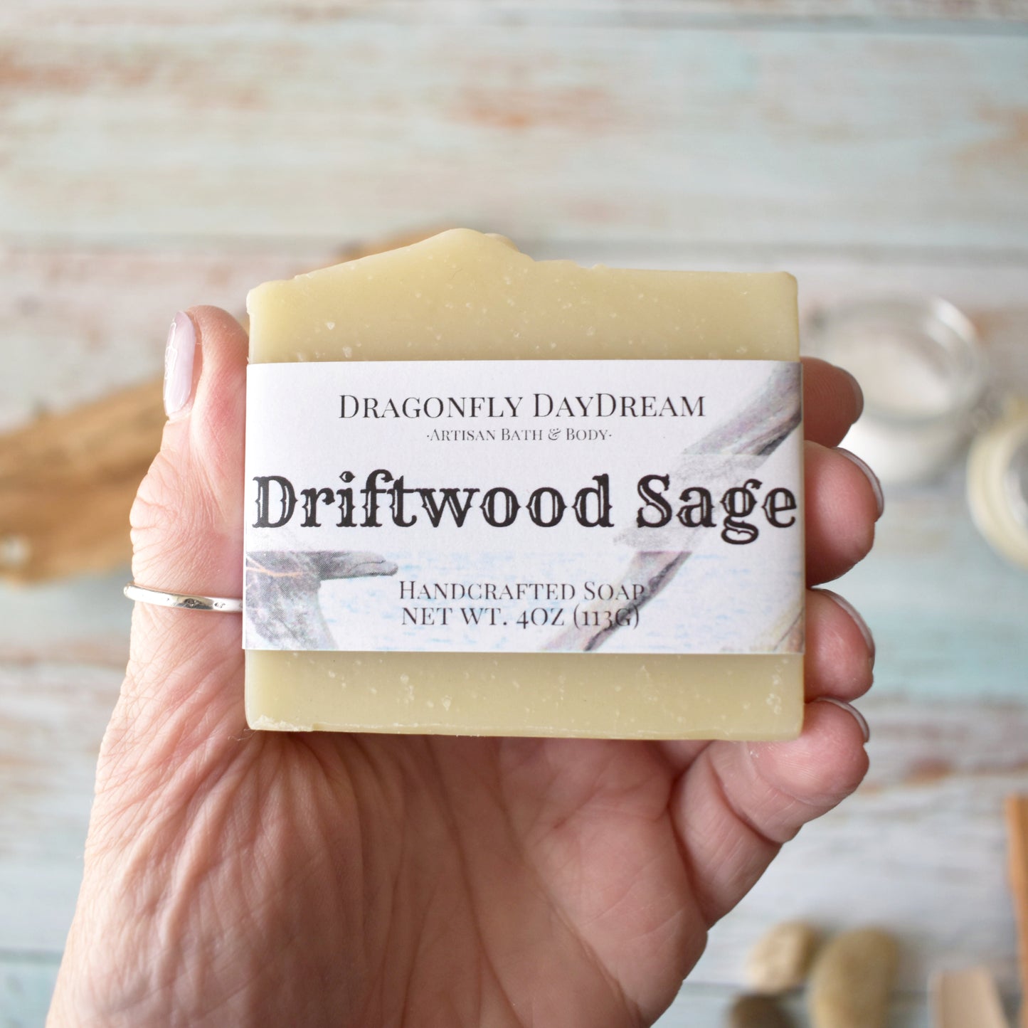 Rectangular beige soap labeled “Driftwood Sage, Handcrafted Soap, net wt. 4oz (113g), Dragonfly DayDream, Artisan Bath & Body. Soap is showcased in hand to show size. Background is blurred weathered light blue wood with smooth river stones, driftwood and jar of course sea salt.