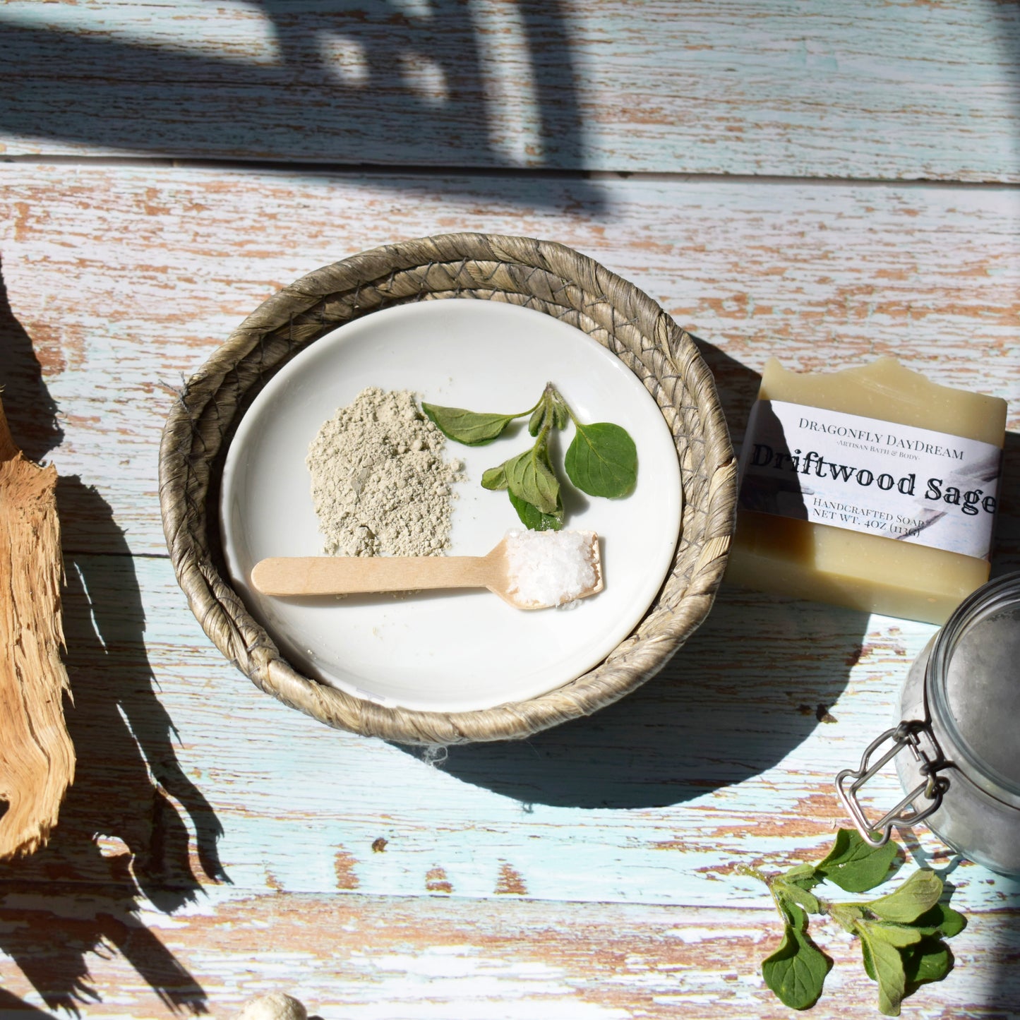 Sunlight from the left. A white plate in a rattan bowl with three element of the plate: a small pile of fine ground dry french green clay, a little pit of greenery, and a small bamboo spoon with a scoop of course white sea salt. To the top right of the plate is a labeled rectangular beige soap, to the left is a small piece of driftwood with shadow.