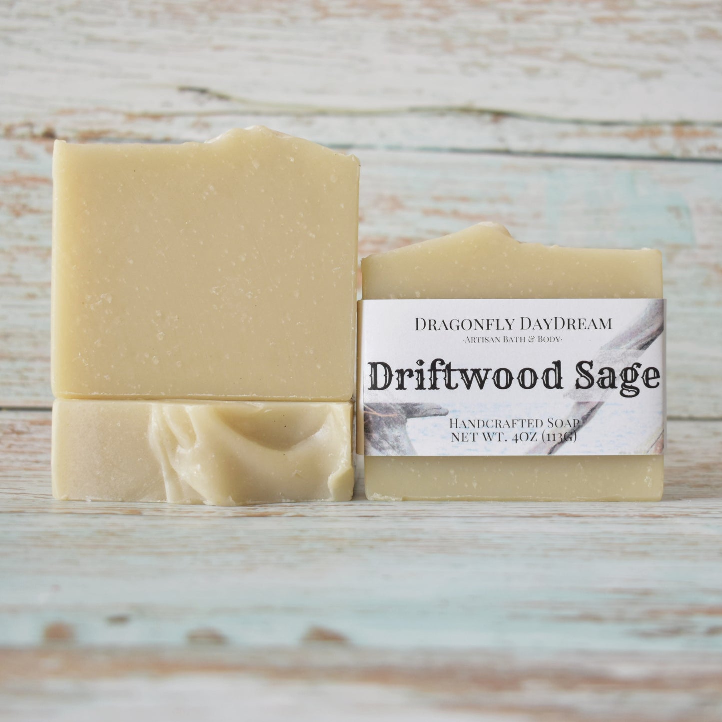 3 rectangular beige soaps. One soap is labeled “Driftwood Sage, Dragonfly DayDream, Artisan Bath & Body, Handcrafted Soap, Net Wt 4oz (113g)”. Weathered light blue wood background