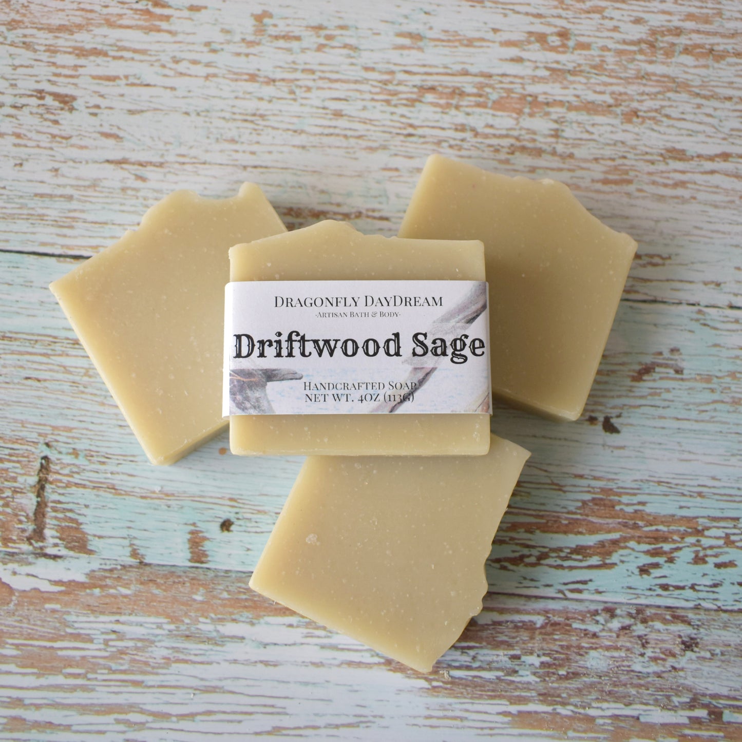 4 rectangular beige soaps sitting on a weathered light blue wood background. 3 soaps are displayed laying down with one labeled soap on top. Label “Driftwood Sage, Handcrafted Soap, Net Wt. 4oz (113g), Dragonfly DayDream, Artisan Bath & Body:.