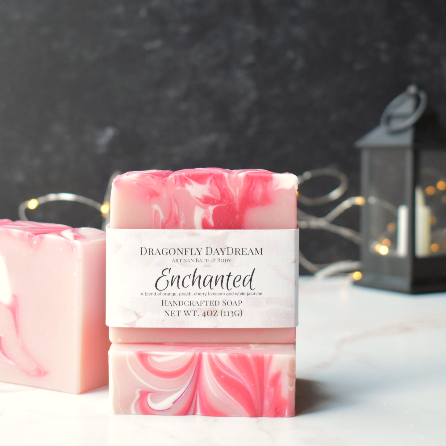 Pink, white and dark pink swirl soap on a marble counter with a dark charcoal background. Rectangular soap in front is stacked on another and labeled “Dragonfly DayDream, Artisan Bath & Body, Enchanted, A blend of orange, peach, cherry blossom and white jasmine, Handcrafted Soap, Net Wt. 4oz (113g)”. Background is blurred with yellow fairy lights and black lantern
