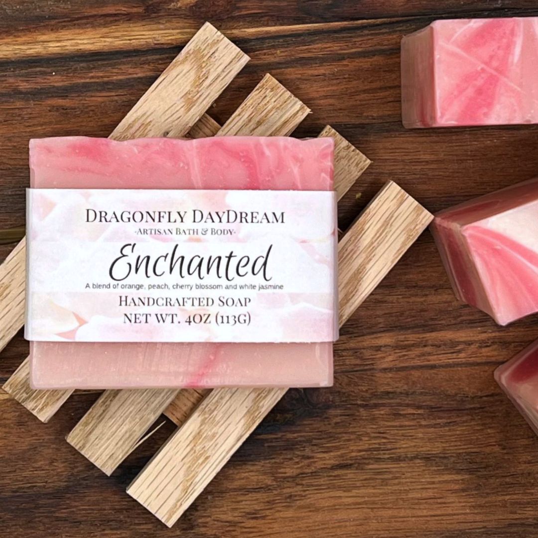 One labeled pink swirled bar of soap. Label “Enchanted, A blend of orange, peach, cherry blossom and white jasmine, Handcrafted Soap, Net Wt. 4oz (113g), Dragonfly DayDream, Artisan Bath & Body. Soap is sitting on slatted wooden soap tray on top of a dark brown wood table. To the right is the top view of three light and dark pink swirled soaps
