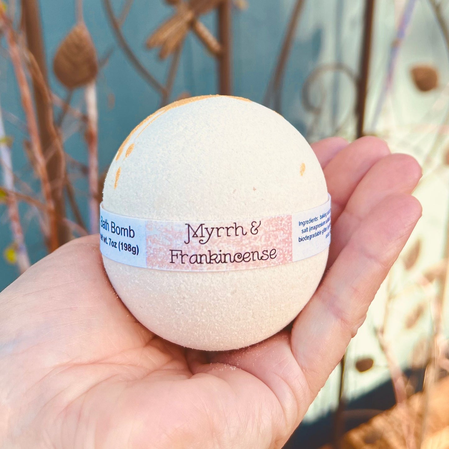 Light beige 7oz bath bomb with a wrap around label “Myrrh & Frankincense” set in a hand to showcase size. Blurred background is taken outside agains a dark turquoise background and wire plant stand with dragonfly.