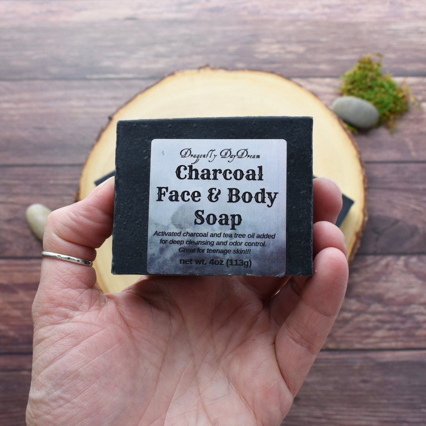 Charcoal Face & Body Soap Bar that is labeled “Activated Charcoal and Tea Tree oil added for deep cleansing and odor control. Great for teenage skin. Soap is shown in a hand to show size. The background is a medium brown table top with a tree round, stone and moss.
