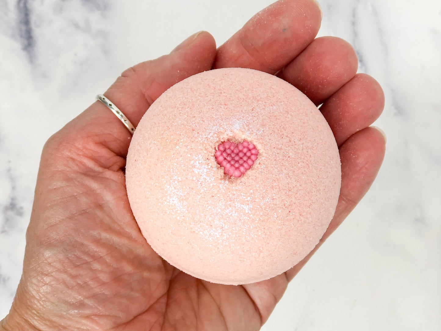 Light pink/peach round bath bomb with mini candy heart and iridescent glitter on palm of hand to show scale. Marble countertop in background