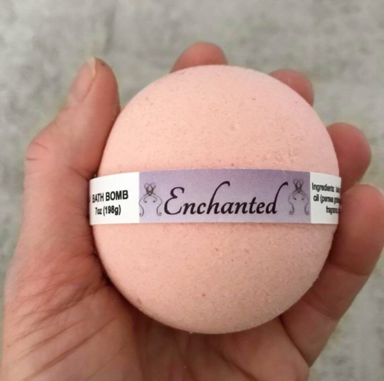 Round light pink/peach labeled bath bomb on palm of hand. Grey background