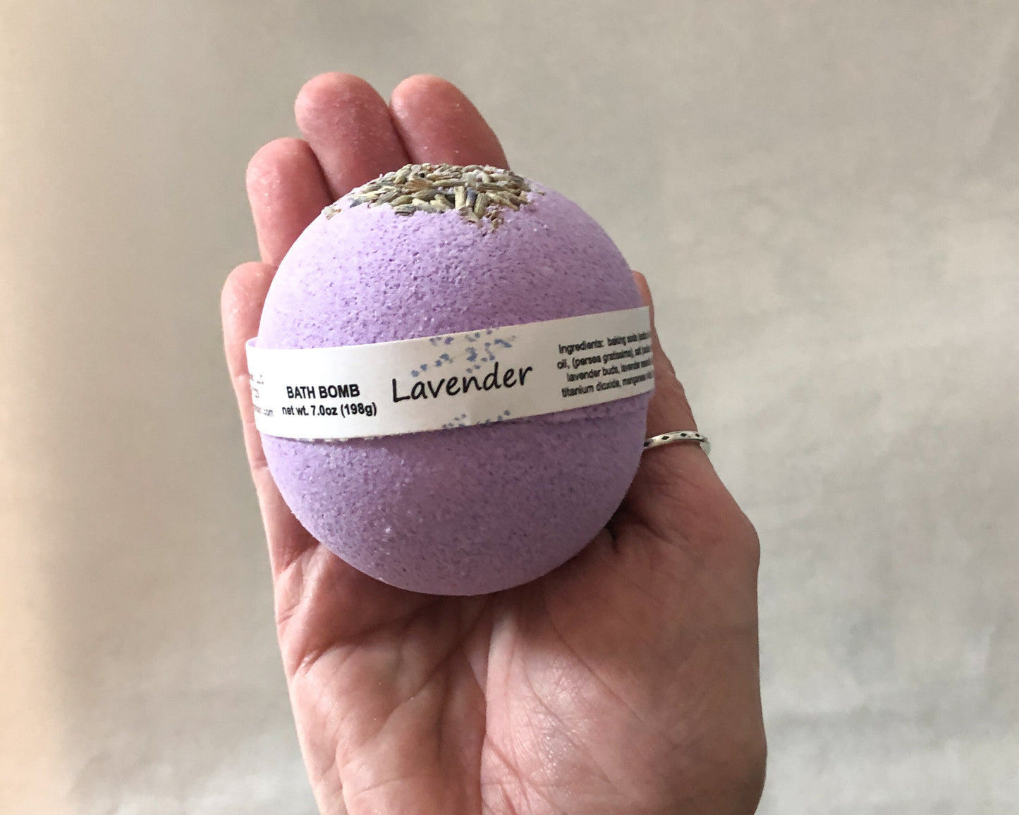 Purple round bath bomb with lavender buds on top with label on hand for scale. Background is white towel.