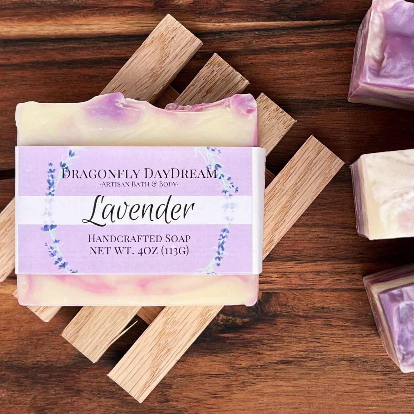 Lavender Handcrafted Soap, Dragonfly DayDream, Artisan Bath & BOdy, Net Wt. 4oz (113g). Soap bar has lavender color swirls in a beige base. Soap bar sits atop a wooden slatted soap tray on a dark brown table top. There are 3 Lavender soap bars off to the right