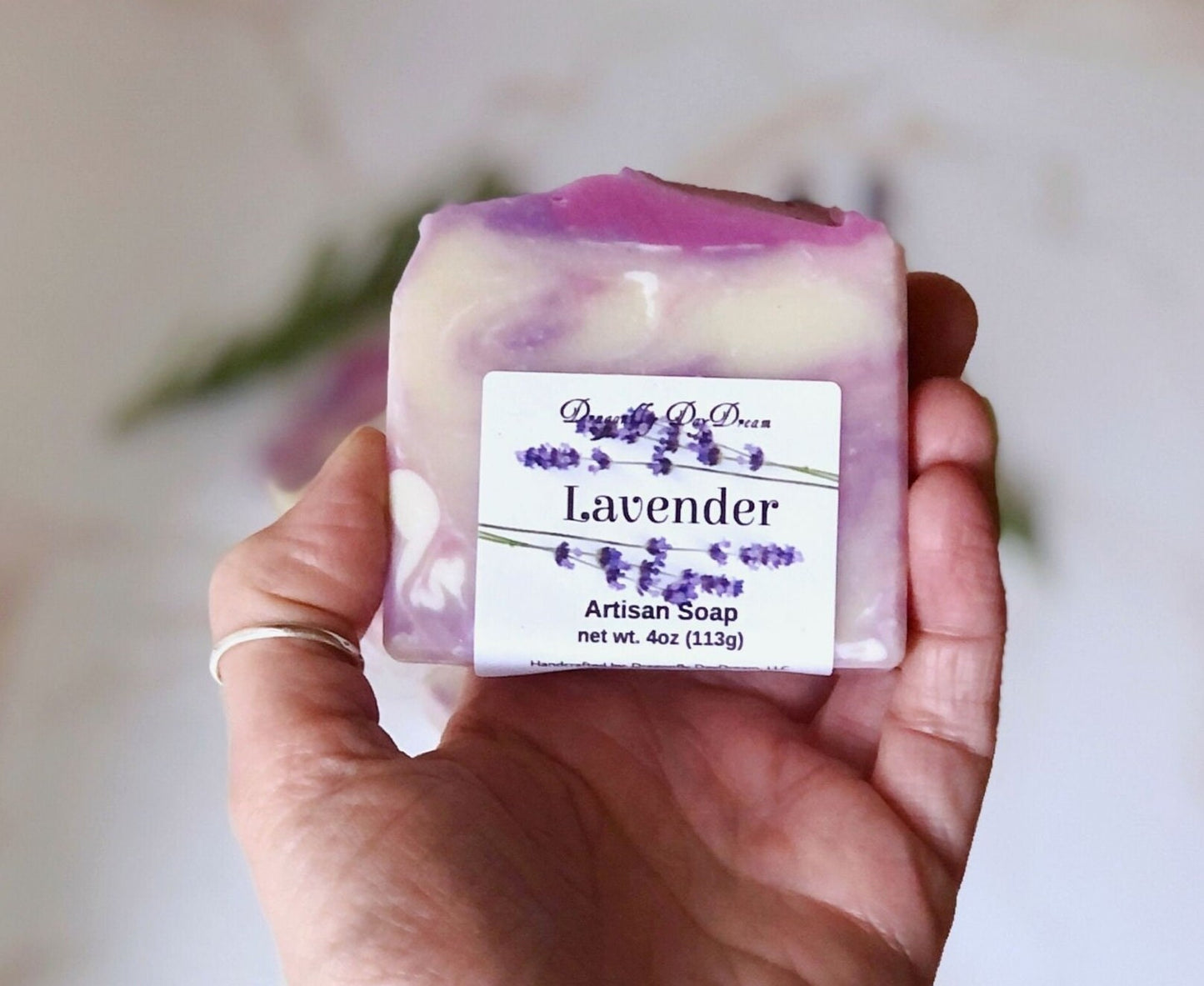 Label: Lavender Artisan Soap, Dragonfly DayDream, net. Wt. 4oz (113g), rectangular soap with light lavender and darker purple swirls held in a hand. Background is blurred purple soap and greenery