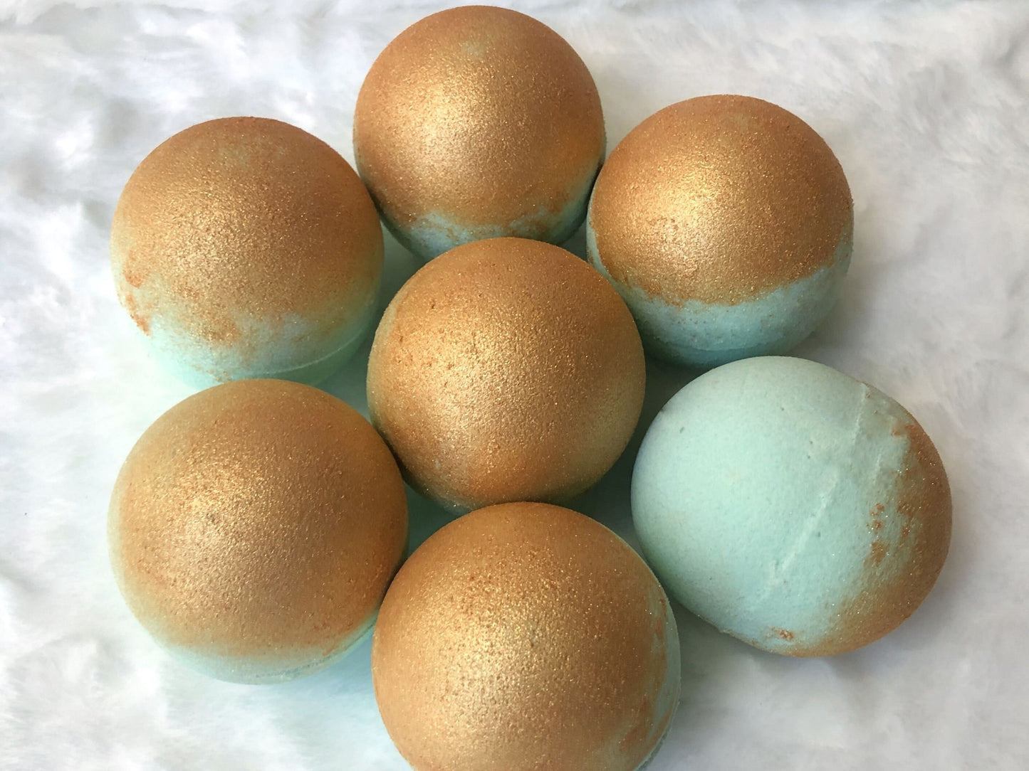 7 Light teal bath bombs sitting on a fuzzy white background. Bath bombs have golden mica on half of bath bomb