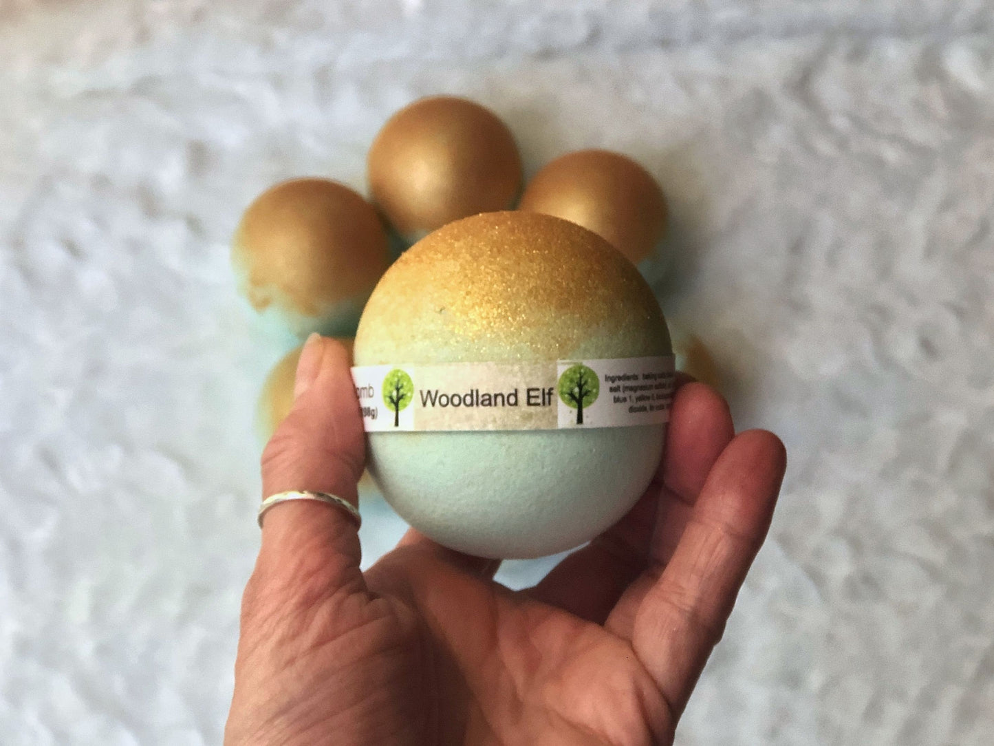 Light Teal Bath bomb with label and gold mica on top. More gold and teal bath bombs in background on white fuzzy material.