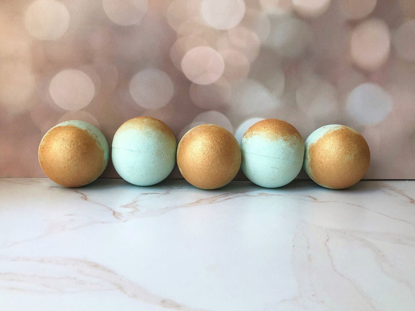 5 large 7.8oz bath bombs lined up in a row. Bath bombs are light teal with gold mica on top sitting on a marble countertop with a blurred fairy light background
