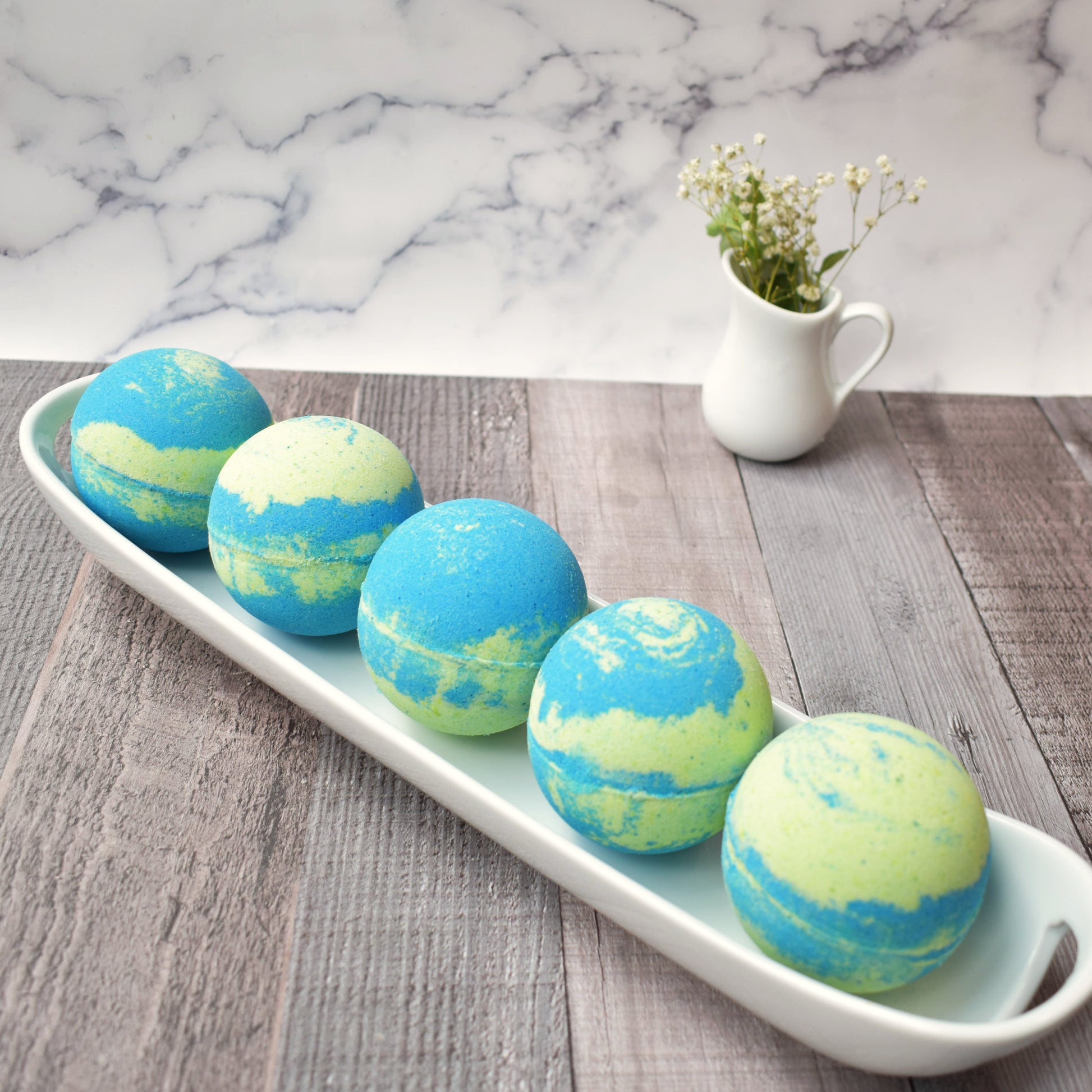 5 Green & Blue swirled bath bombs on a white tray.  Small white creamer carafe with baby’s breath are in background Sitting on a light brown/grey wood table and marble backboard