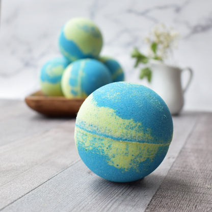 Close up of a Lime green and blue swirled bath bomb.  In the background are 4 bath bombs stacked on a square wood tray. The the right is a small white creamer carafe with baby’s breath flowers.  All sitting on a wood brown/grey table and marble background