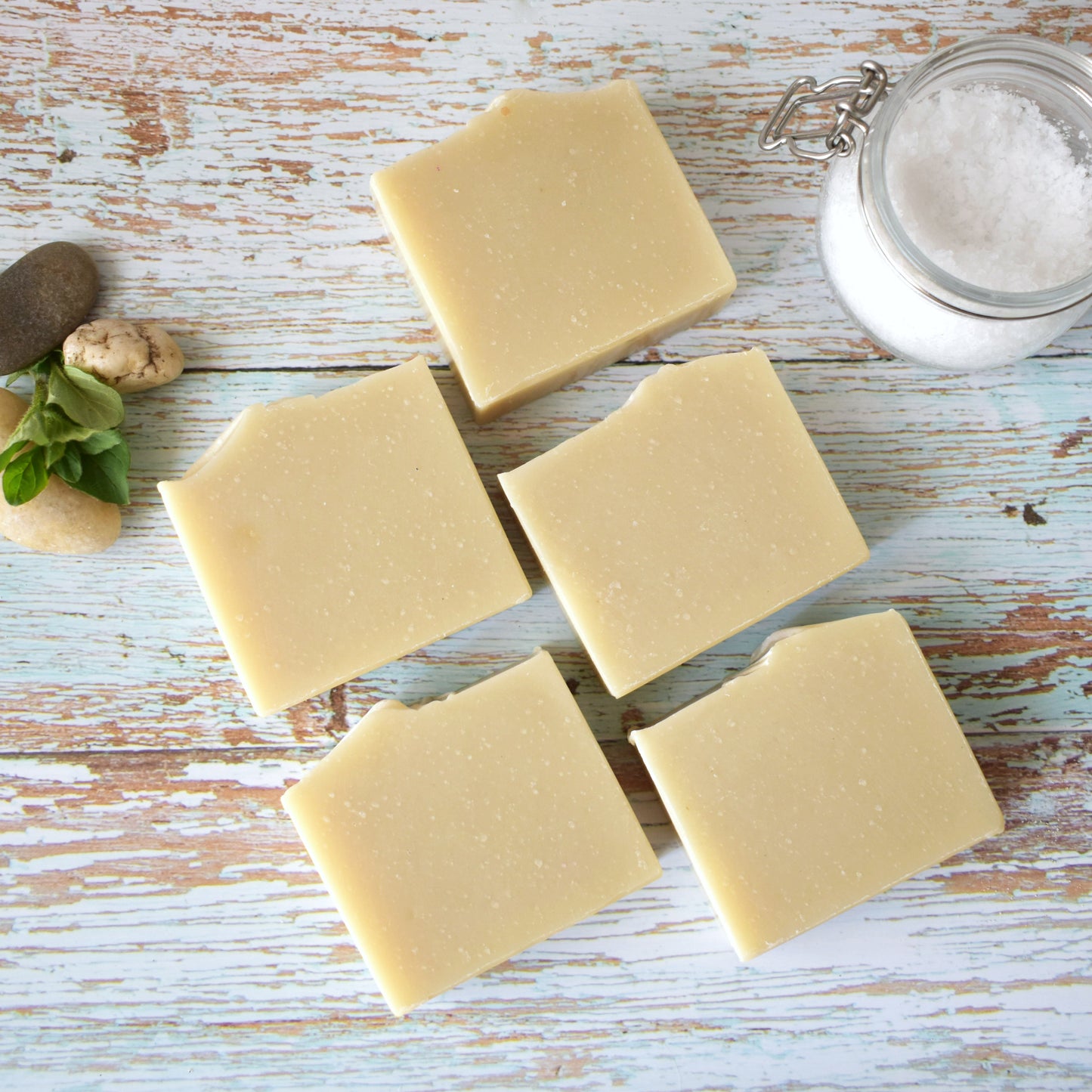 5 rectangular beige soaps laying in two rows on a weathered light blue wooden background. To the upper right there is a small glass jar with course sea salt and to the upper left are 3 small river stones.