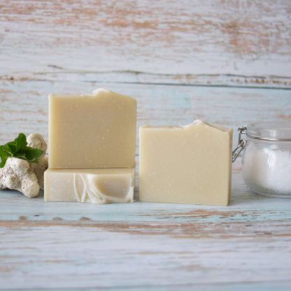 3 beige soaps sitting on a weathered light blue wood background.  To the left of the soap is white coral with greenery and to the left is a lightening sealed jar with course salt.