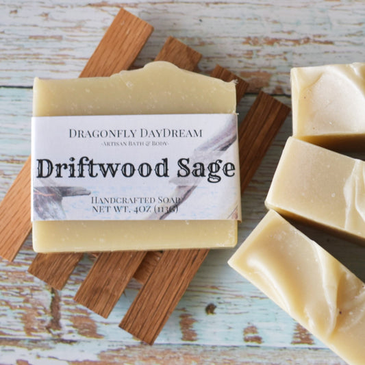 Driftwood Sage Artisan Soap Bar sitting on top of a slatted wooden soap tray.  To the right are 3 additional soaps with a blueish weathered wood background