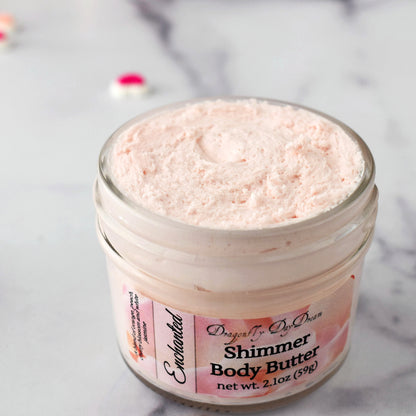 One glass jar of shimmer body butter opened to show label and light pink whipped body butter on a marble background with a couple of pink candy hearts in background