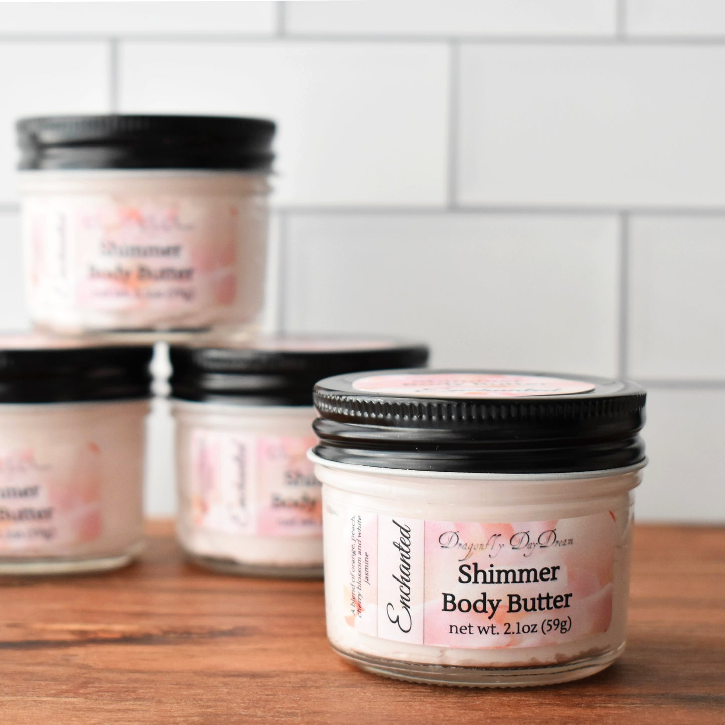 1 jar of shimmer body butter in the foreground with black lid and labeled “Enchanted Shimmer Body Butter, Dragonfly DayDream, net wt. 2.1oz (59g)” on a medium brown wood table and a white subway tile background.  Also in background are 3 jars of body butter out of focus.
