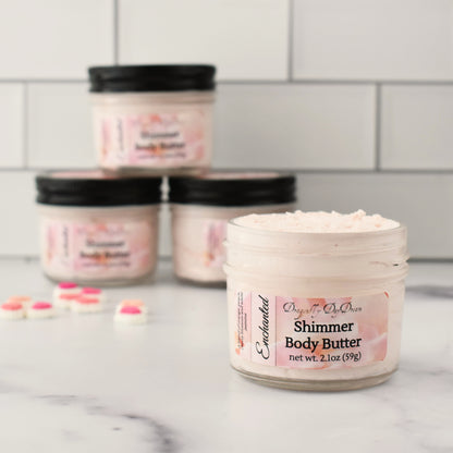 One jar of body butter in foreground labeled “Enchanted, Dragonfly DayDream, Shimmer Body Butter, net wt. 2.1oz (59g)”, showing light pink body butter in jar on a marble countertop and a white subway tile in background.  Blurred background of 3 jars of whipped body butter and a handful of pink candy hearts