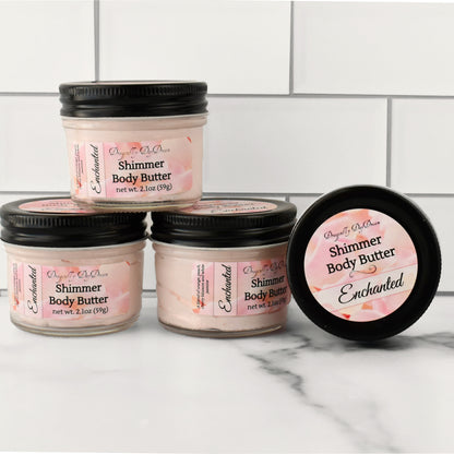 4 jars of shimmer body butter on a marble countertop with a white subway tile background.  One jar is on it’s side showing the top label “Dragonfly DayDream, Shimmer Body Butter, Enchanted”.  The other 3 jars are stacked and show front label with same writing plus “net wt. 2.1oa (59).
