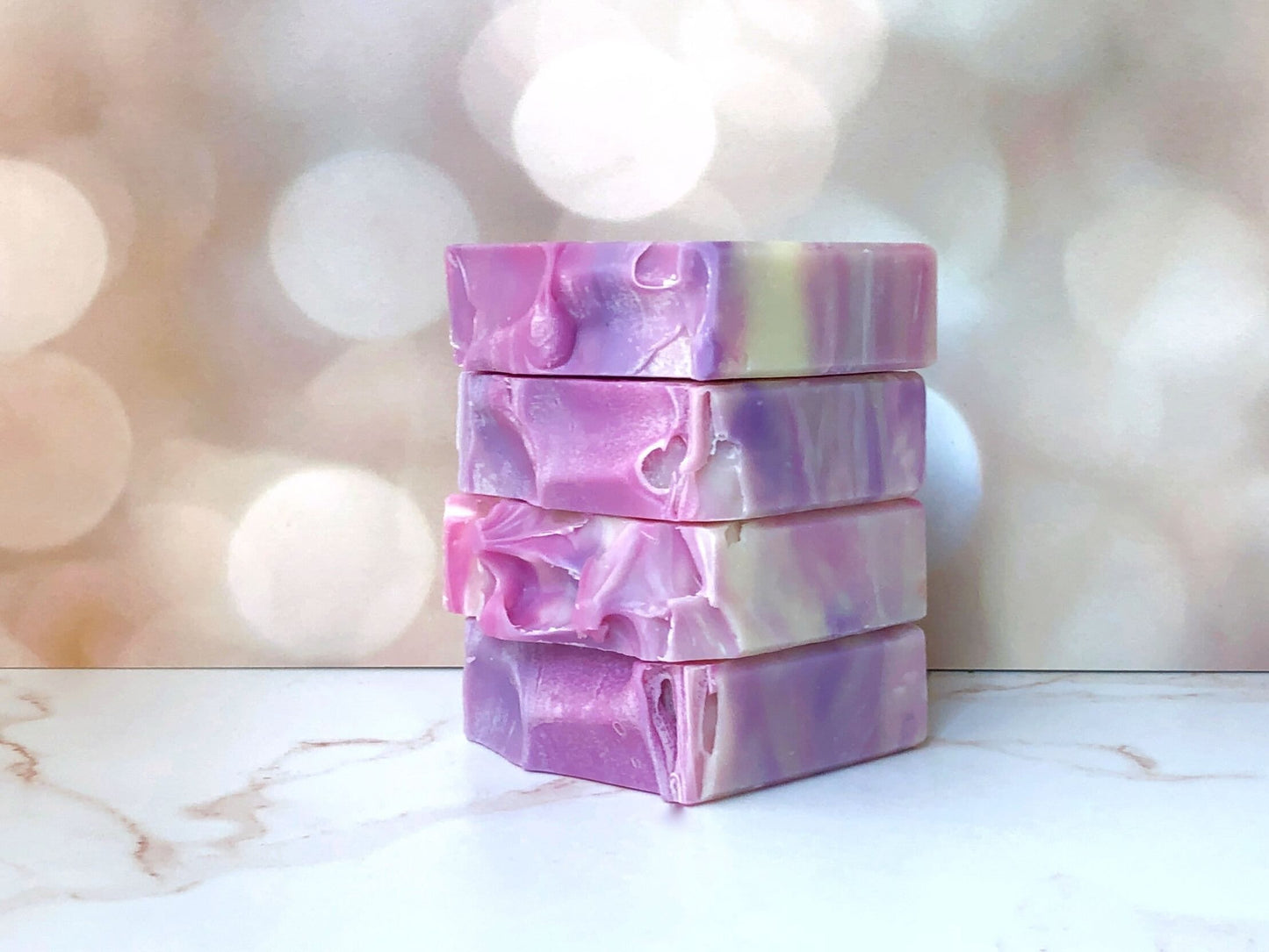 Stack of 4 rectangular soap bars sitting atop a marble countertop and a blurred fairy light background.  Soap is lavender and grape purple color swirled in a beige base.