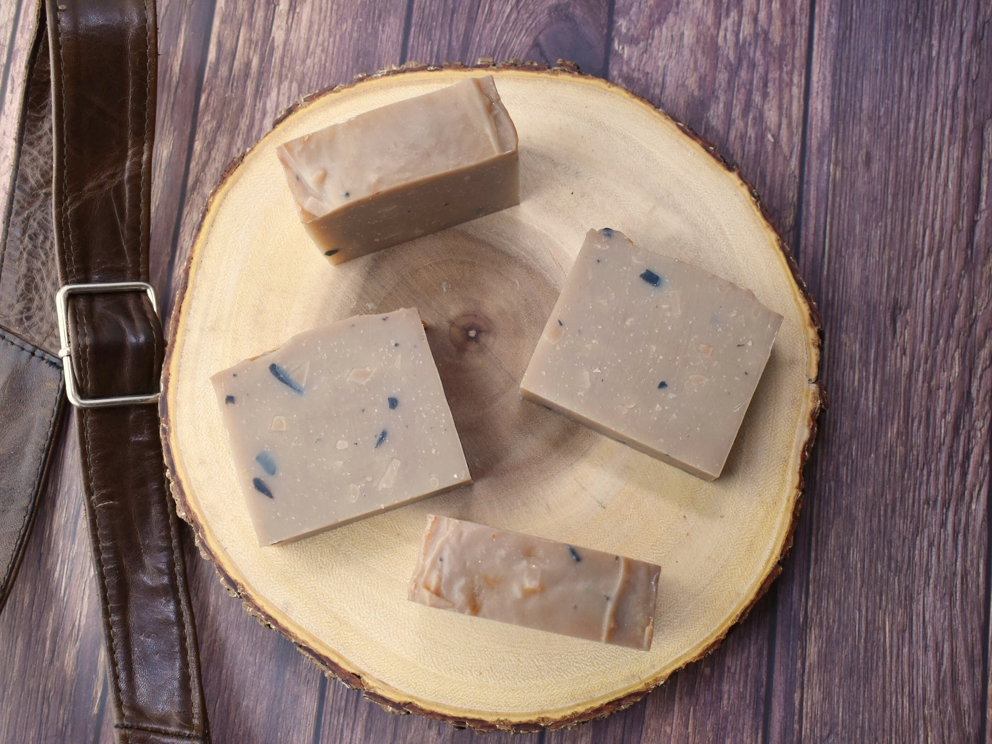 Photo shows 4 Leather Artisan Soaps, two laying down and two standing.  Soaps are featured on a wood round and leather strap next to 