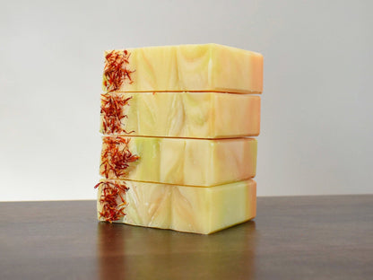 Stack of 4 Lemongrass Dreams Artisan Soap.  Soap is yellow, orange, with streaks of lime green.  Bright orange safflower petals lay atop of the soap on one side.  Soap is sitting on a dark brown wood table.