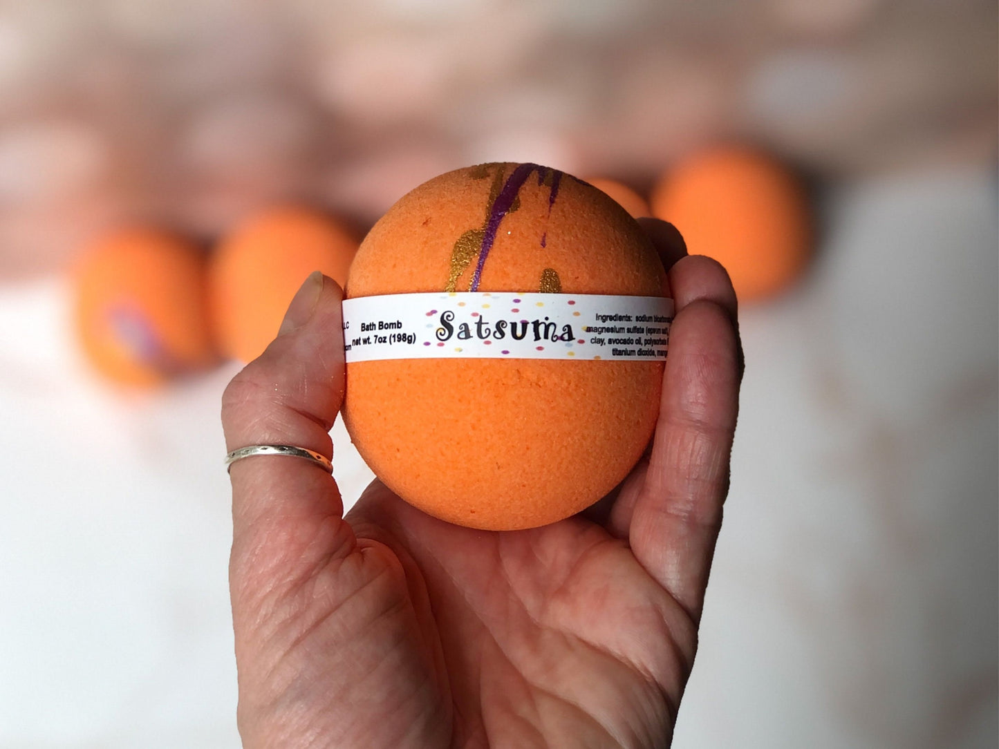 Satsuma Bath Bomb 7.8oz labeled with ingredients and “bath bomb” handheld.  Bath Bomb is bright orange with dark purple and gold splashes of color on top.  Background is blurred with 4 satsuma bath bombs sitting on a marble countertop