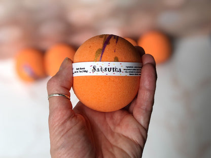 Satsuma Bath Bomb 7.8oz labeled with ingredients and “bath bomb” handheld.  Bath Bomb is bright orange with dark purple and gold splashes of color on top.  Background is blurred with 4 satsuma bath bombs sitting on a marble countertop