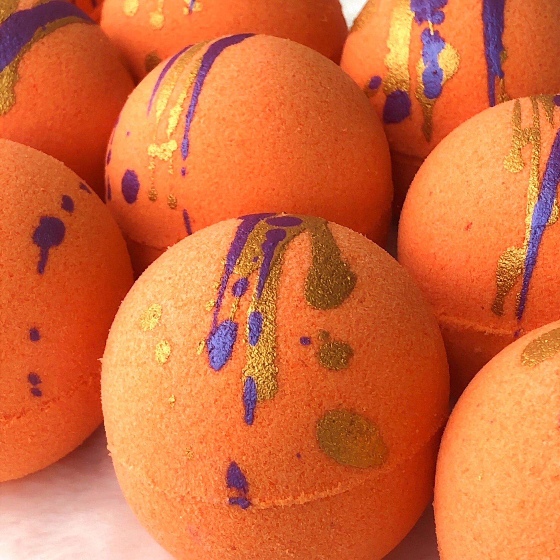 Bright orange satsuma bath bombs with dark purple and gold “splashes” of color.