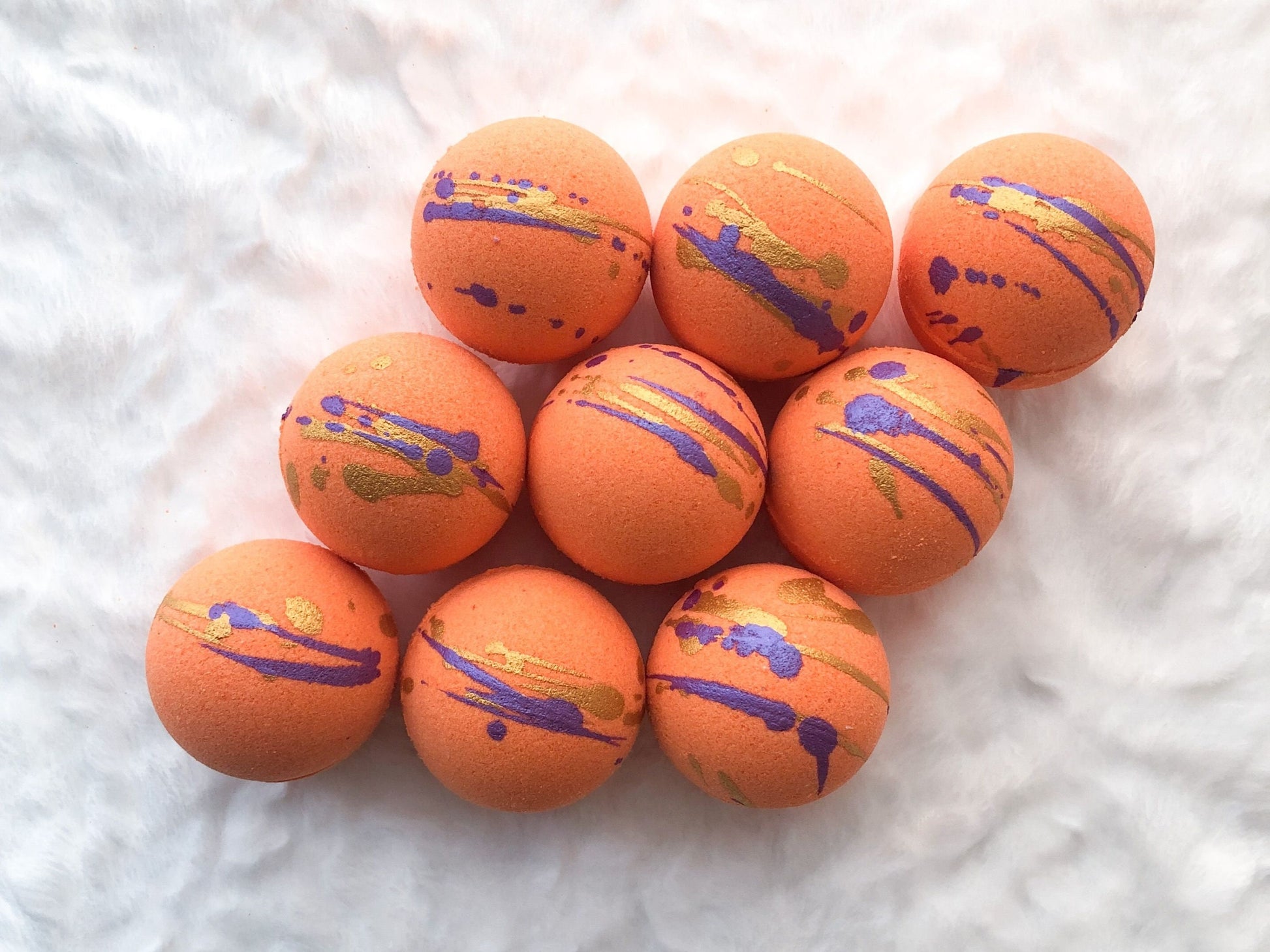 Top view of 9 bright orange Satsuma bath bombs which have dark purple and gold splashes on top.  Background is a fuzzy white blanket.