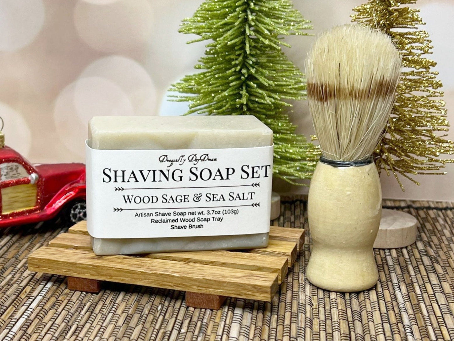 Wood sage and sea salt soap on a reclaimed wood soap tray with a shave brush beside it. Background features blurred fairy lights, two sparkly trees, and a red truck ornament.