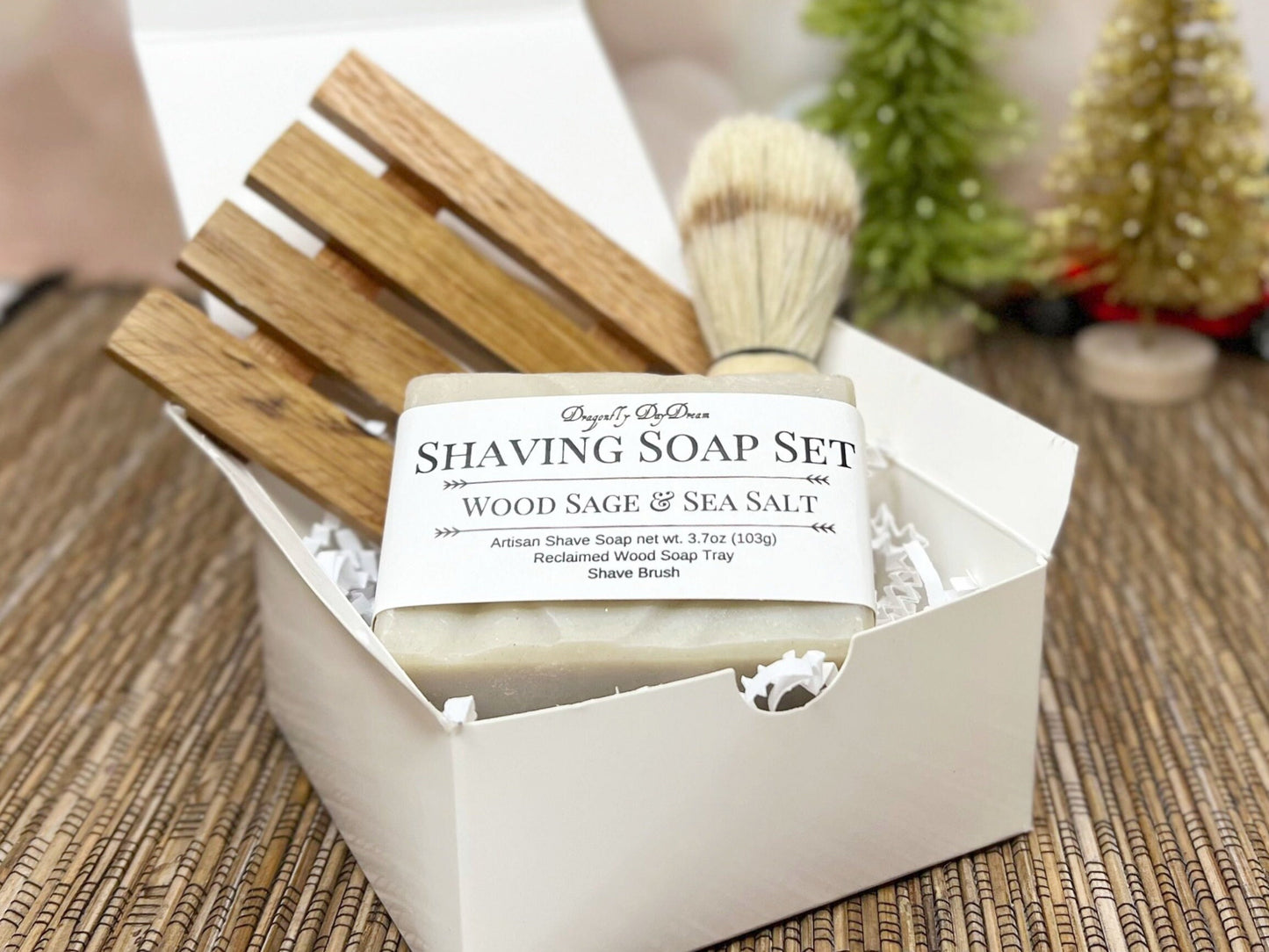 White gift box with one bar of wood sage & Sea salt shave soap, along with a reclaimed wood soap tray and shaving brush.  Gift box is sitting on a straw mat on a countertop with glittery green and gold trees in background