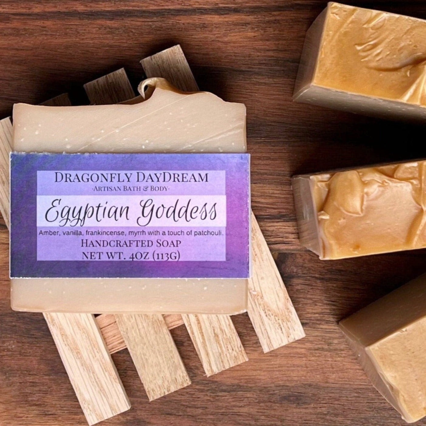 Tan Egyptian Goddess soap labeled with a purple tag, resting on a recycled wood soap tray atop a dark brown table. To the right, three additional Egyptian Goddess soaps are displayed from a top view.