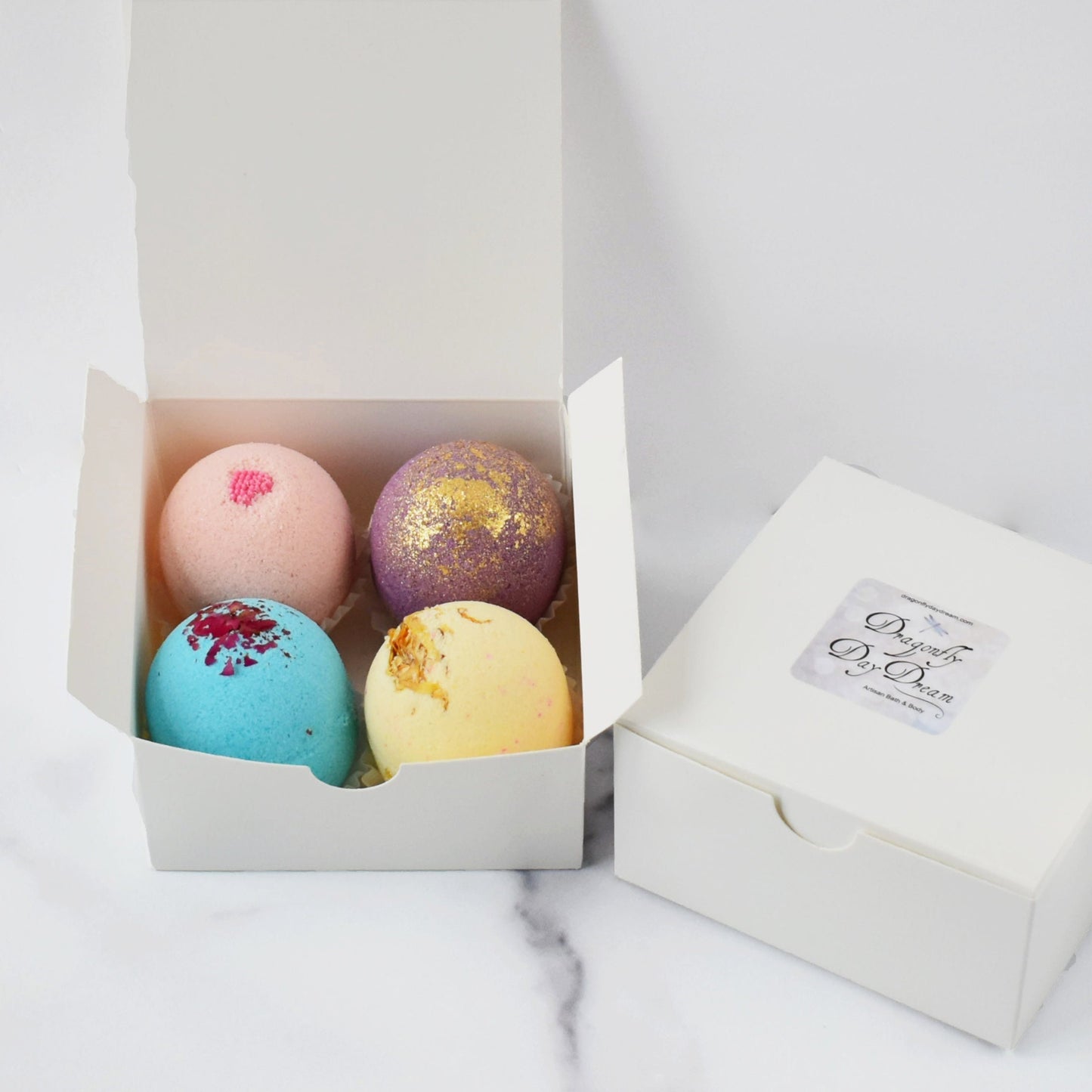 Four mini bath bombs in pink, blue, yellow, and purple colors arranged in a white gift box, with a branded white gift box beside it, all placed on top of a marble background.