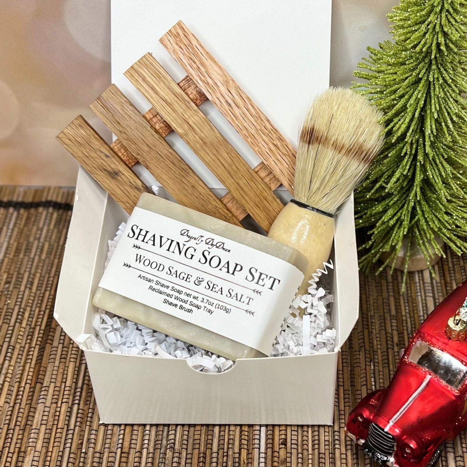 White gift box with wood sage and sea salt soap, reclaimed wood tray, and shaving brush on grass mat, with sparkly tree, red truck ornament, and blurred fairy lights in background.